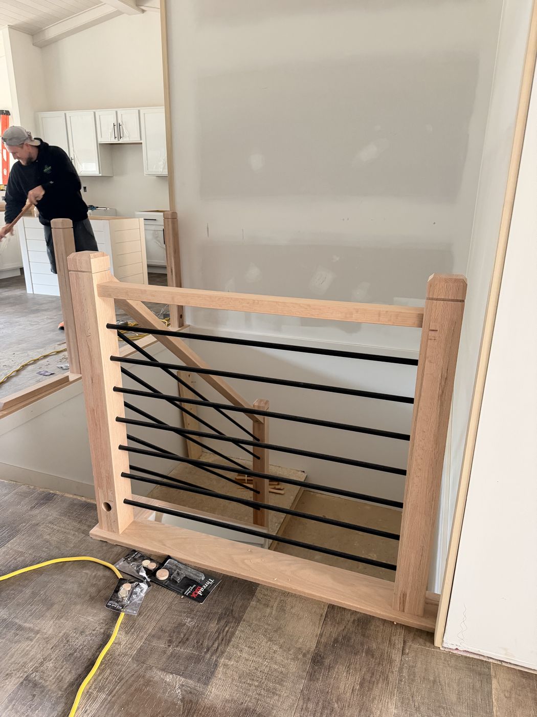 Before: Raw wood stair rail with horizontal black bars dry-fit, with Dakota Robinson sweeping behind it